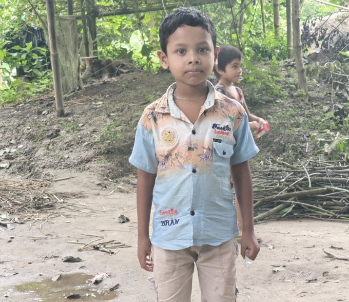 A young boy stands in the foreground wearing a blue and beige patterned shirt featuring "BRAUM" and a smiley face graphic, with another child visible in the background amid a rural outdoor setting with branches and greenery. The image has no direct connection to the cryptocurrency-focused post about "$ANSEM," which appears unrelated to the rural village scene depicted.