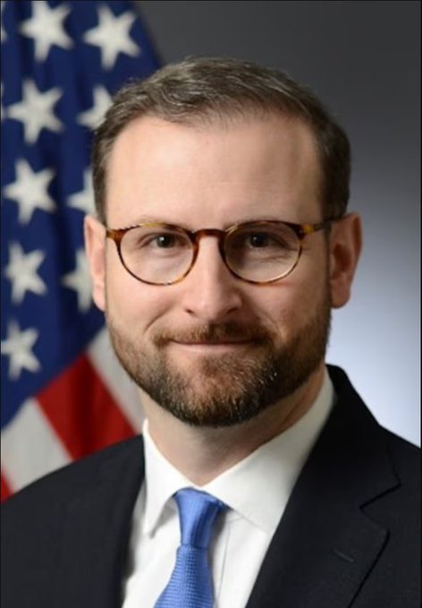 An official portrait of a man with glasses, a beard, and a blue tie in front of an American flag.