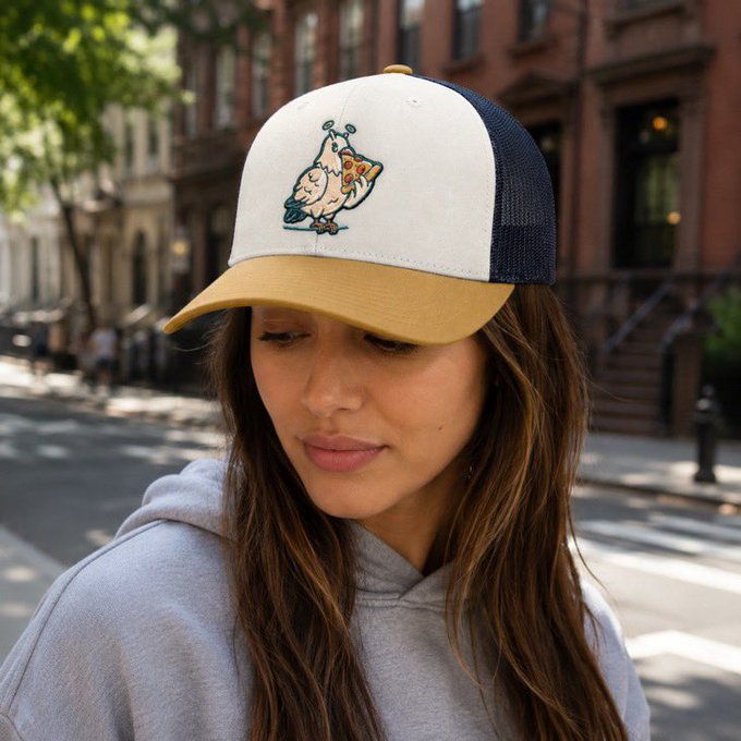A woman with long brown hair wearing a gray hoodie and a two-tone trucker hat featuring an embroidered cartoon chicken holding a slice of pizza, standing on a city street.