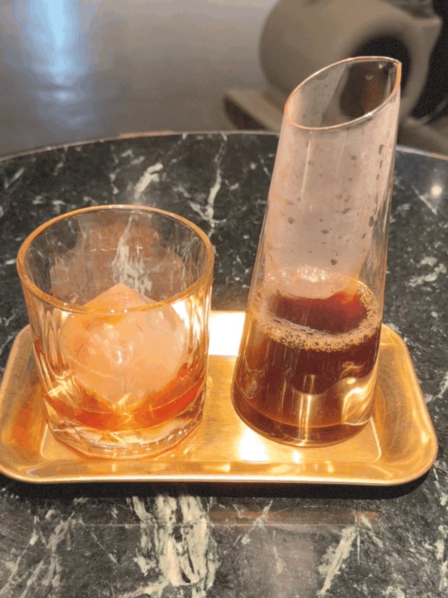 The image features two glasses of liquid sitting on a tray placed on top of a marble table. One glass has ice cubes in it, while the other contains a beverage. The scene appears to be set up for a social gathering or event where people can enjoy drinks and conversation together.