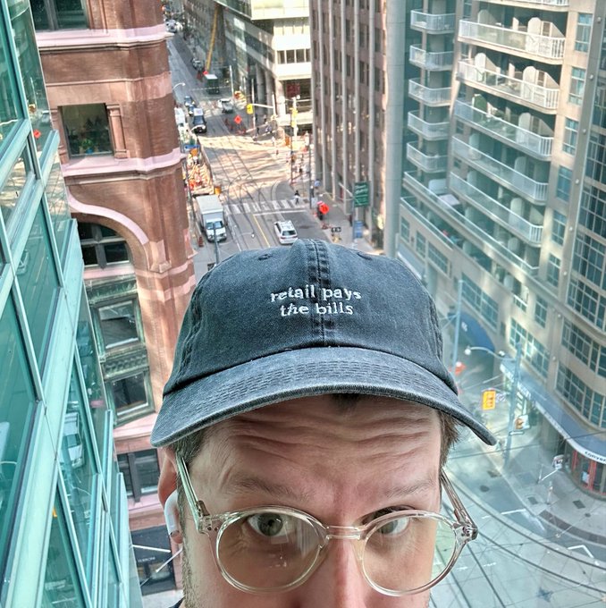 A man wearing clear-framed glasses and a dark baseball cap reading "retail pays the bills" takes a selfie from a high-rise overlooking a bustling city street, visually embodying the post's message that retail investors are the driving force behind HBAR's potential growth and adoption.