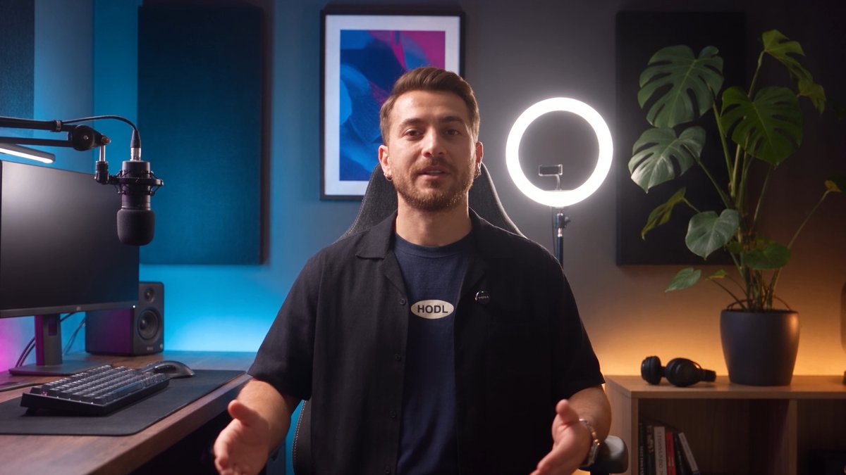 A man wearing a dark shirt with an oval "HODL" badge speaks in a home studio, gesturing with both hands, while surrounded by a professional microphone, ring light, framed blue artwork, and a monstera plant, illustrating a tutorial on depositing and withdrawing USD via BtcTurk.