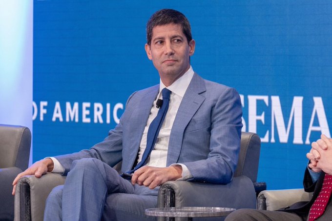 A man in a blue suit and tie sits on stage at what appears to be a banking conference, with 'BANK OF AMERICA' partially visible in the background.