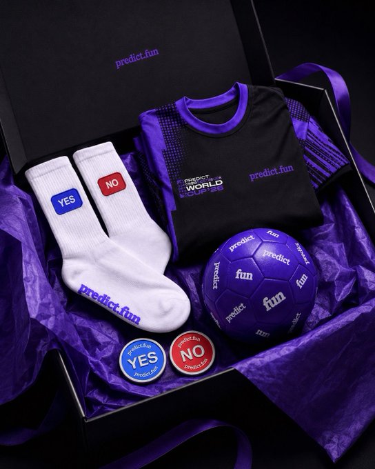 The image features a gift box containing three sets of soccer-themed merchandise, including a soccer ball, two pairs of socks with "No" written on them, and a purple soccer jersey. The items are neatly arranged inside the box, which is placed against a black background. The text in the image reads "Focus on what is in the image and how it relates to predict! @predictdotfun The World Cup prediction event has kicked off— which team will you pick to win the championship?".