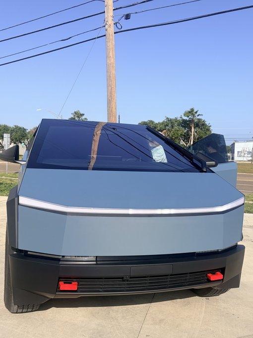 The front view of a silver Tesla Cybertruck parked on a street under power lines and a clear blue sky.