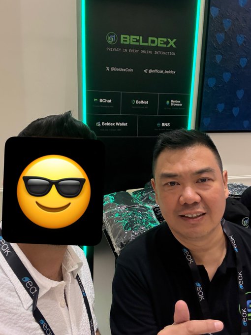 Two men pose for a photo at a Beldex booth with promotional banners reading 'BELDEX - PRIVACY IN EVERY ONLINE INTERACTION' and listing services like BChat, BelNet, and Beldex Wallet; one person's face is covered by a sunglasses emoji.