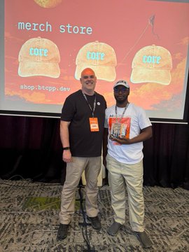 Two men pose in front of a "merch store" screen displaying "COME" hats and a BTC shop link (shop.btcppl.com), with one holding a book titled "21 Lessons." The image reflects the post's message by capturing a moment at an educational Bitcoin workshop, showcasing the next generation of miners learning about the technology.