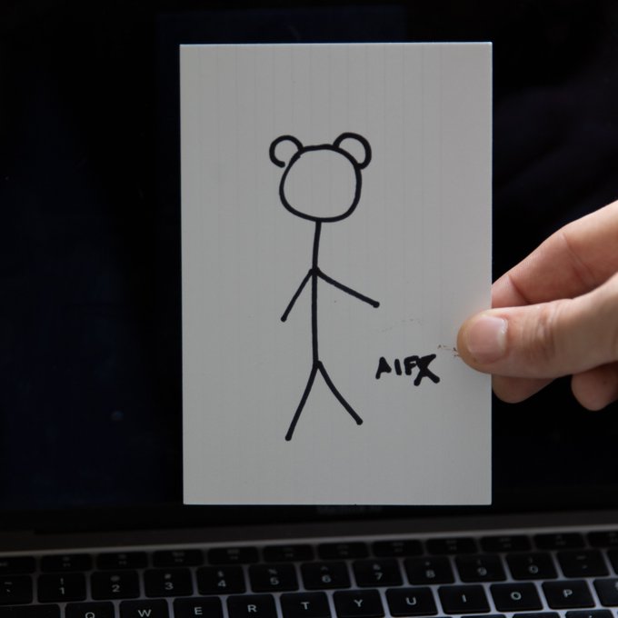 A hand holds up a white card featuring a simple stick figure drawing with two round ears and the handwritten text "AIFX" beside it, photographed in front of an open laptop keyboard.