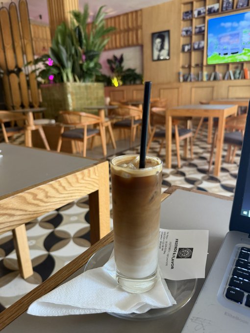 A tall iced coffee with milk sits on a napkin at a Starbucks-style cafe with wooden furniture and patterned tile floors, next to a laptop with a 'Patron Flavor' coffee sample card.