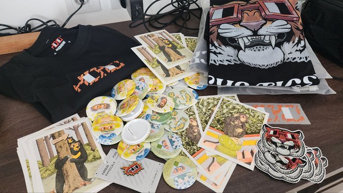 Merchandise from a "Nouns x AthenaX" collaboration—including t-shirts, stickers, and branded coasters and cards featuring the Nouns character—spread out on a table, ready for distribution at CoinfestAsia.