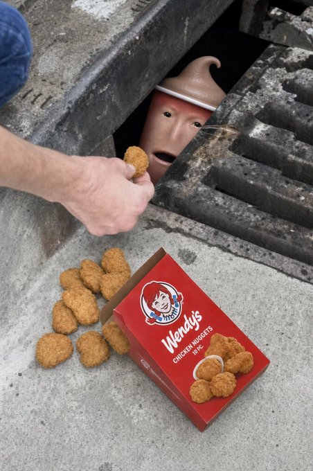 A person's hand is feeding Wendy's chicken nuggets to a creepy mannequin head wearing a pointy hat, which is peering out from a storm drain next to an open box of Wendy's nuggets.
