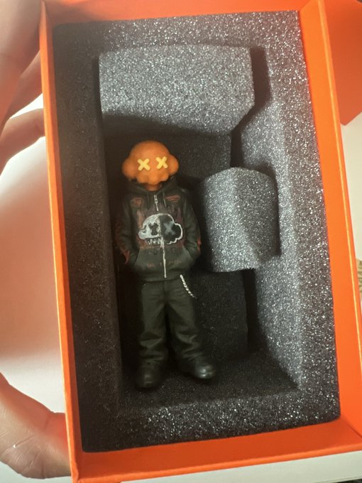 A small designer toy figure with an orange skull-like head marked with 'X X' eyes sits in black foam packaging inside an orange box, wearing a black hoodie with a skull design.