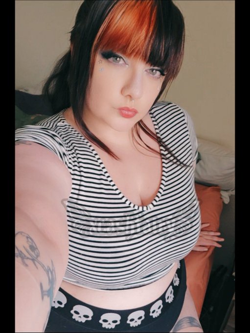 A young woman with dark hair and orange highlights poses for a selfie, wearing a black and white striped crop top and black shorts with skull patterns.
