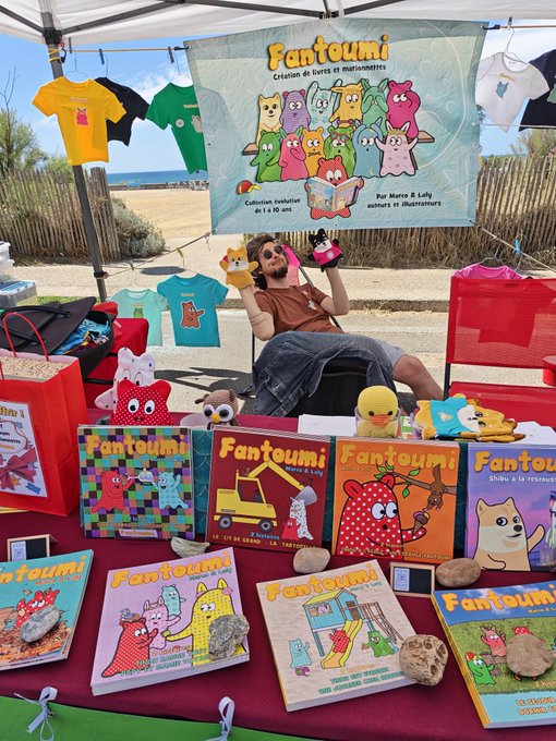 An outdoor vendor stall called 'Fantoumi' displaying children's books and plush toys at a beachside market, with a man relaxing in a chair behind the table.