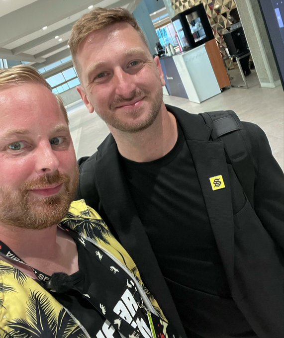 Two men pose for a selfie, one wearing a tropical print shirt and the other in a black blazer with a yellow SXSW badge, taken in what appears to be a convention center hallway.