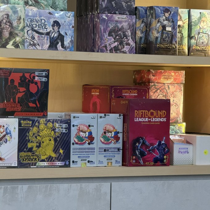 A shelf display at a store featuring Pokémon Trading Card Game products, including Grand Archive, Riftbound: League of Legends, and PanTheon booster boxes and elite trainer boxes.