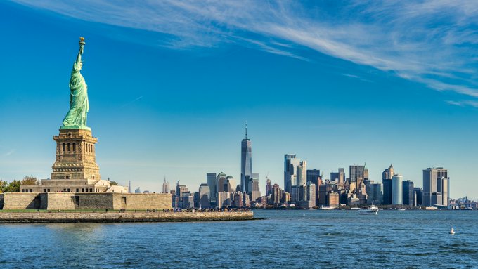 The Statue of Liberty stands on the left with the New York City skyline, including One World Trade Center, visible in the background across the water.
