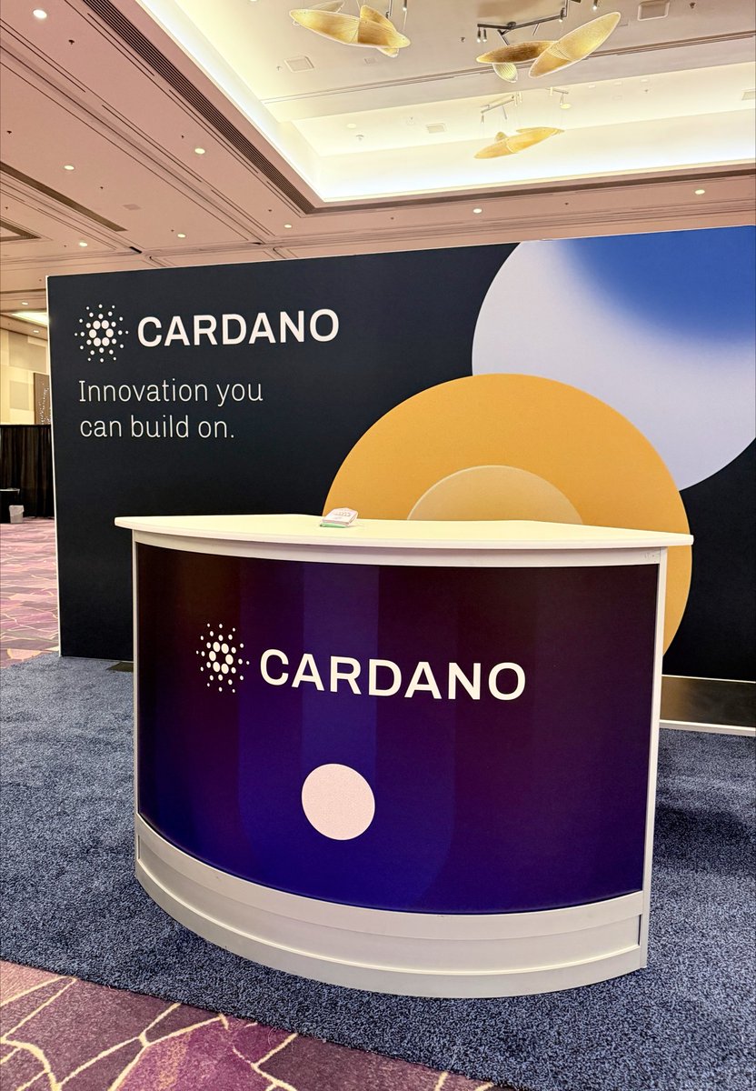 A trade show booth or reception area featuring a purple counter and large backdrop with the 'CARDANO' logo and the tagline 'Innovation you can build on.' displayed in a hotel ballroom setting.