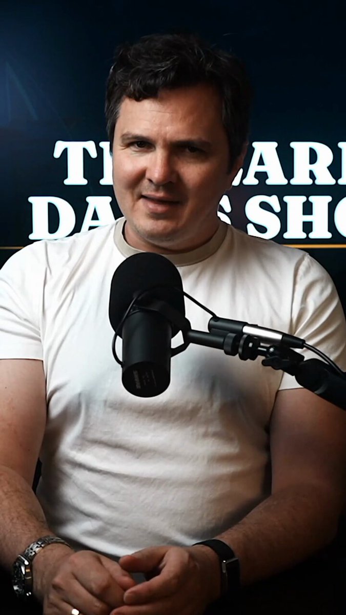 The image features a man wearing a white shirt, sitting behind a microphone with his hands folded in front of him. He appears to be posing for a photo or recording a podcast interview. The background includes a sign that reads "The Darjeeling Show" along with an image of a clock and the words "Recency bias is why you keep on selling your Bitcoin." This suggests that the man might be discussing the concept of recency bias in relation to cryptocurrency, as it is a common cognitive error where people tend to rely more on recent information or experiences when making decisions.