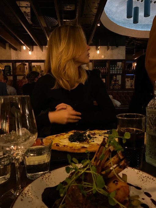 A blonde woman in a black top sits at a restaurant table with a pizza and drinks in the foreground, under a decorative ceiling fixture.
