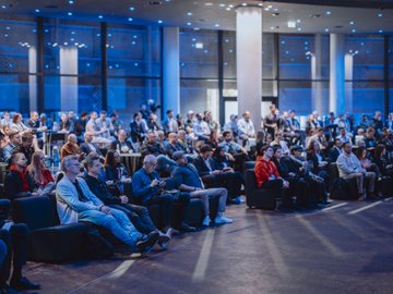 A large audience attends a conference event bathed in blue lighting, listening attentively in a modern venue with floor-to-ceiling windows. The image captures the global, in-person nature of the gathering referenced in the caption, illustrating the diverse attendees who came together across cities—from Berlin to Nairobi—to discuss blockchain's enterprise and public sector applications.