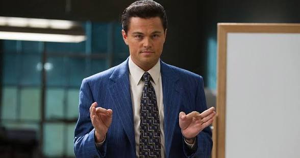 Actor Leonardo DiCaprio in a blue pinstripe suit and patterned tie, gesturing with his hands while standing near a whiteboard, in a scene that appears to be from a film.