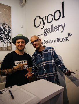 Two men posing together inside Cycol Gallery by Solana x BONK, with one wearing a hat and Bowery shirt, and a guestbook visible on the counter.