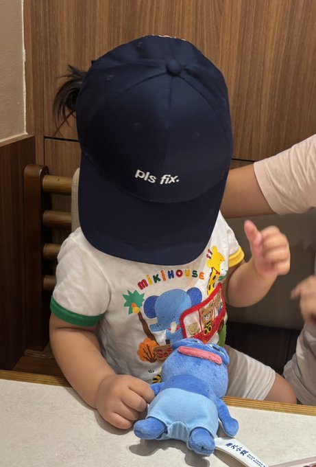 A young child wearing an oversized navy blue "pls fx." cap and a Mickey Mouse t-shirt sits at a table holding a small blue Stitch plush toy.