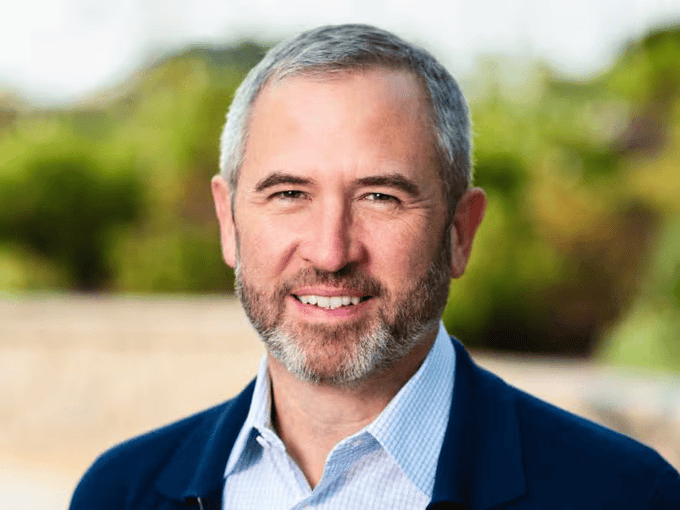 The image features a man wearing a blue jacket, smiling at the camera with his eyes closed. The caption reads "TODAY: Ripple CEO Brad Garlinghouse says Jamie Dimon'S pushback on the Clarity Act is intentional misrepresentation," which seems unrelated to the main subject of the photo and could be an unrelated comment or statement about the image itself.