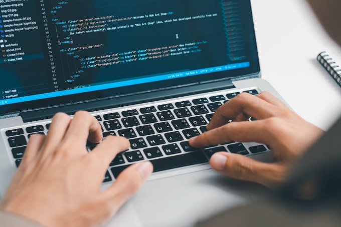 A close-up of hands typing on a laptop keyboard with HTML code visible on the screen, referencing 'HBA Art Shop' and 'Kaos49' shop link classes.