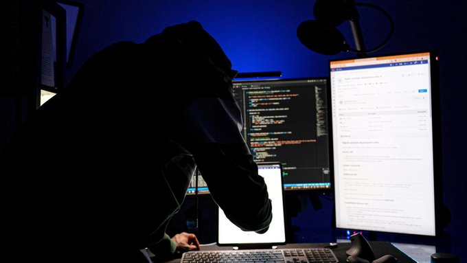 Dark silhouette of a hooded person working on multiple computer monitors displaying code and data in a dimly lit room with blue lighting, evoking a hacker or programmer.