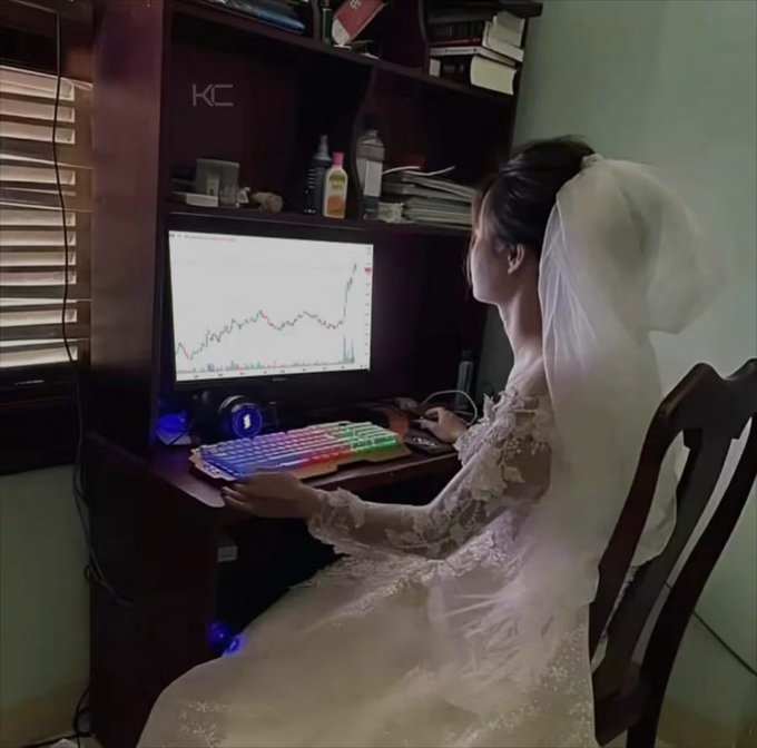 A bride in a white wedding gown and veil sits at a computer desk looking at a stock chart with a sharp upward trend on the monitor, humorously suggesting that her partner's financial market success is a sign she "chose the right one." The image playfully captures the idea of marrying well, both literally and in terms of investments.