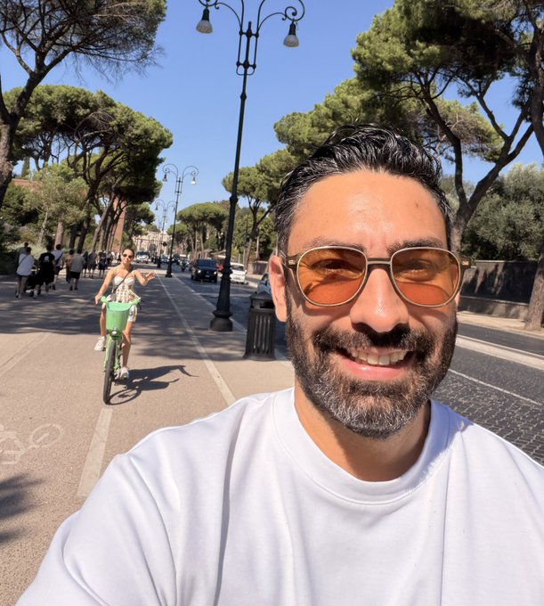 A smiling bearded man wearing sunglasses takes a selfie on a tree-lined street while a woman rides a green bike in the background, suggesting a sunny day in Rome.