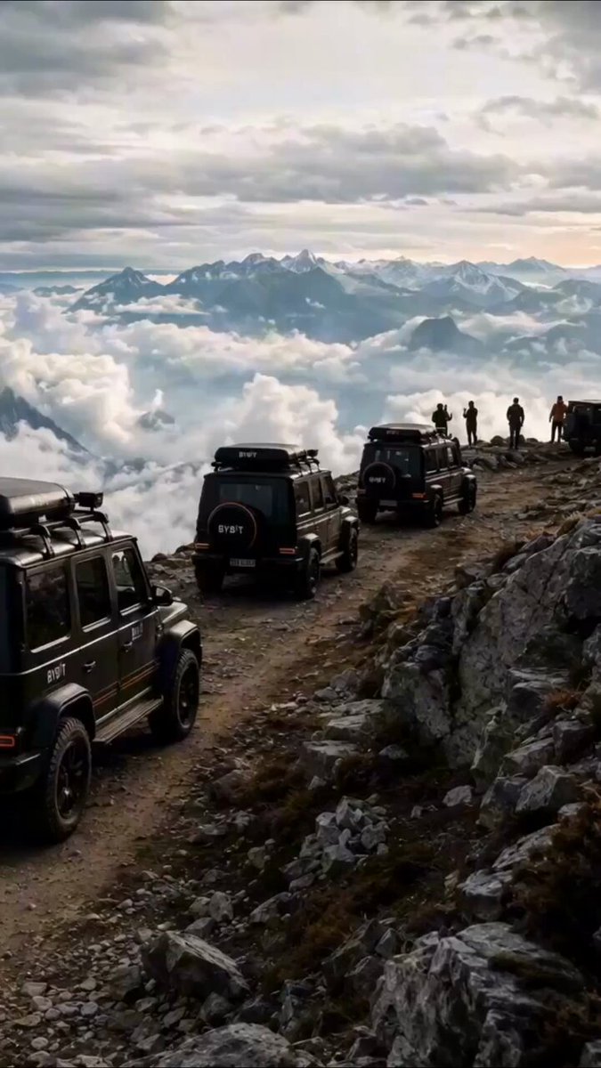 The image shows a group of people standing at the top of a mountain, overlooking a valley below with clouds. There are several cars parked nearby, including some black jeeps. The scene suggests that these individuals have come together for an adventure or outdoor activity in this picturesque location. The presence of the cars and jeeps indicates that they might be on a road trip or exploring the area, possibly seeking out scenic spots like the one depicted in the image.