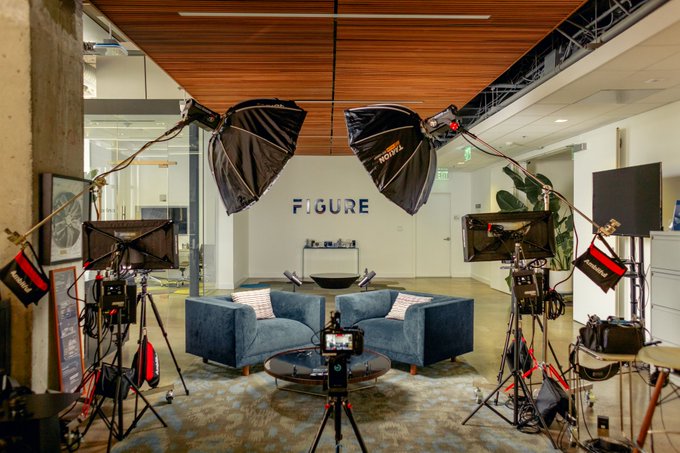 An indoor photography or video studio setup at the 'FIGURE' office, featuring blue armchairs, professional lighting equipment including softboxes, and multiple cameras on tripods.