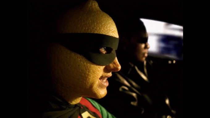 A close-up profile shot of a person wearing a textured yellow mask and a black domino mask, with another masked individual visible in the blurred background.