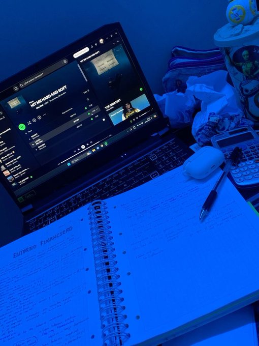A desk scene bathed in blue light shows a laptop open to a music player playing 'HIT ME HARD AND SOFT' by Billie Eilish, alongside a notebook titled 'EMPIRICAL EVIDENCE', a pen, earbuds, a calculator, and a phone.
