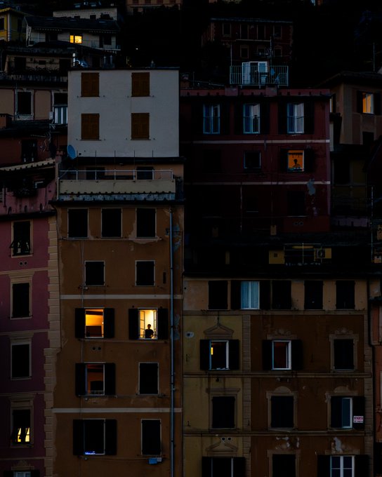A nighttime photograph of stacked, aged apartment buildings with warm light glowing from several windows against the dark facades.