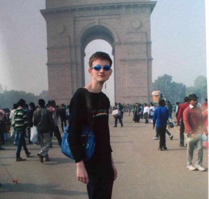 A young man wearing sunglasses and a black long-sleeve shirt stands in front of what appears to be India Gate in New Delhi.