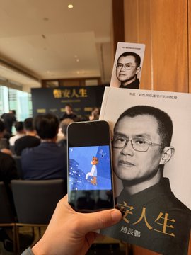 A hand holds up a smartphone displaying an animated character, while simultaneously grasping a large book or brochure featuring a black-and-white portrait of a bespectacled man with Chinese characters reading "定义人生" (Define Life) and "赵长鹏" (Zhao Changpeng/CZ). The setting appears to be a book launch or signing event, with attendees visible in the background near signage. The image's focus on CZ and his memoir "Define Life" connects thematically to the post about NIUMA as a builder community, suggesting the author views the NIUMA/NiuMa project with the same long-term, foundational mindset that CZ embodies in his crypto industry journey, rather than chasing short-term hype.