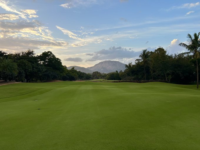 A lush green golf course fairway stretches toward distant mountains under a partly cloudy blue sky, framed by tropical trees on either side.