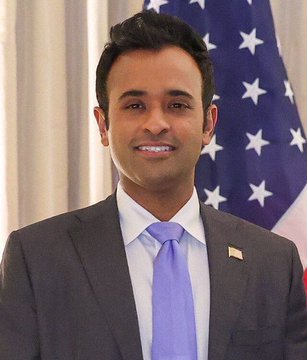 A portrait photograph of Vivek Ramaswamy wearing a gray suit with a lavender tie, smiling and standing in front of an American flag.