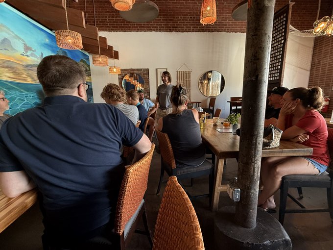 A photograph of a group of people seated at wooden tables in a restaurant or café setting with decorative wicker chairs, hanging lights, and an ocean-themed mural in the background.