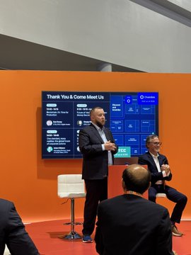 A presentation or panel event with the screen title partially visible, featuring a standing speaker addressing a seated audience in an orange-walled room.