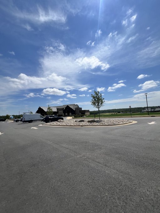 A sunny outdoor view of a parking lot roundabout with a young tree, leading to a building complex under a bright blue sky with scattered clouds.