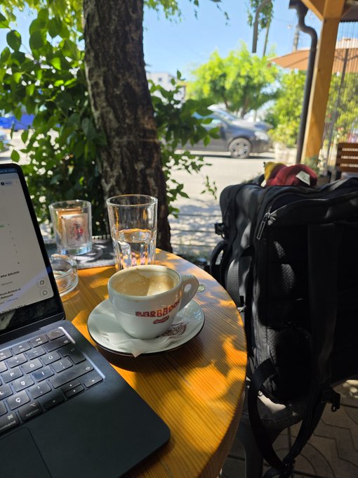 The image shows an outdoor cafe with a wooden table featuring a laptop computer, two glasses of water, and a cup of coffee. A backpack can be seen next to the table, suggesting that someone might have been working or studying there. The scene also includes some potted plants in the background, adding a touch of greenery to the setting. The post about "Gm everyone Working on a nice place" implies that people are enjoying their time at this cafe and possibly taking breaks from work or studies to relax and socialize with others.