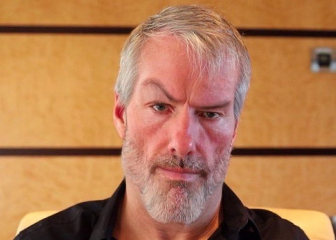 A close-up portrait of a gray-haired, bearded man with a serious expression, set against a wooden panel background — likely Michael Saylor, the executive of Strategy (formerly MicroStrategy). The image pairs with news of Strategy's dramatic financial turnaround, swinging from a $9.9B unrealized loss to approximately $2.1B in unrealized profit.
