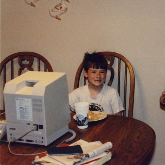 A young boy sits smiling at a wooden dining table with a vintage CRT computer monitor, a cup, and a plate of food, in what appears to be a nostalgic 1990s photograph.