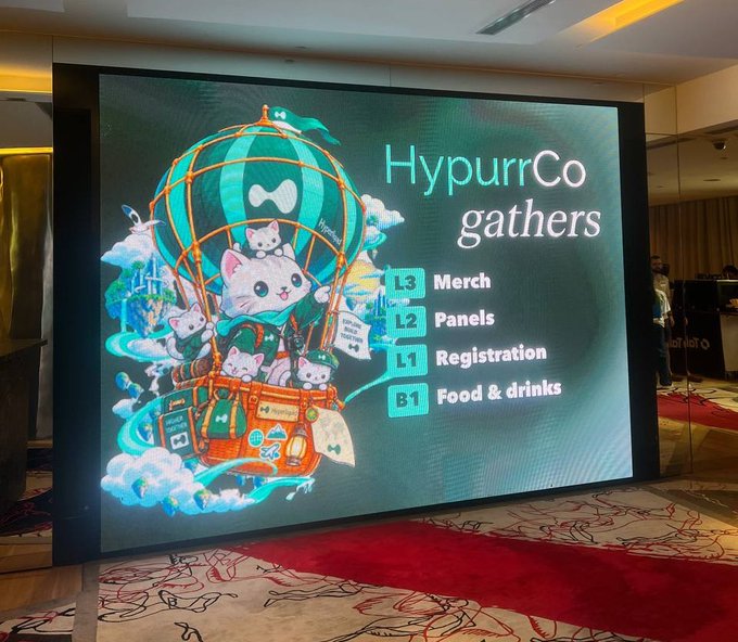 An illuminated event sign for "HypurrCo gathers" displays cartoon cats in a hot air balloon and lists event locations: "L3 Merch," "L2 Panels," "L1 Registration," and "B1 Food & drinks."