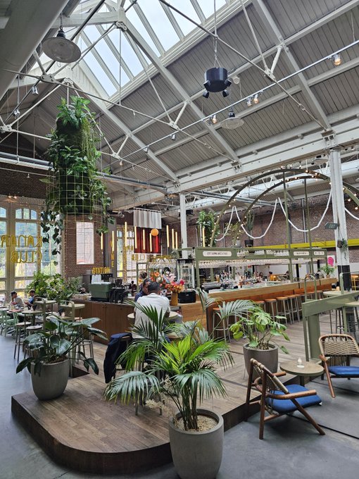 An industrial-style indoor market or food hall with a glass roof, hanging plants, exposed beams, and patrons seated at a bar.